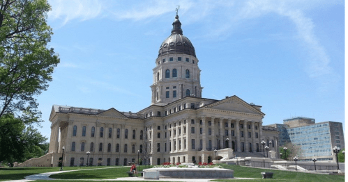 A grand state capitol building with a large dome, surrounded by green lawns and blue skies.