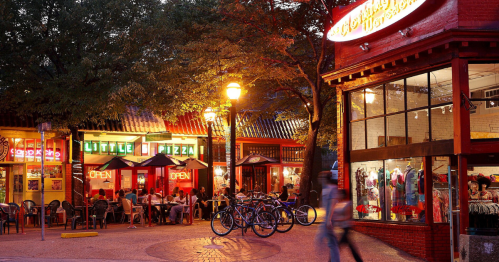 A vibrant street scene at dusk with restaurants, shops, and people enjoying the atmosphere, surrounded by trees and bicycles.