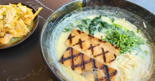 A bowl of creamy ramen topped with grilled tofu and green onions, alongside a small dish of kimchi.