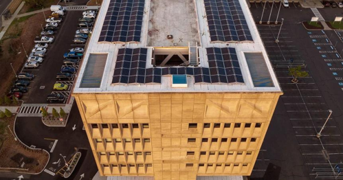 Aerial view of a modern building with a solar panel-covered roof and a large parking lot nearby.