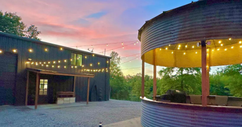 A rustic outdoor space with string lights, a circular bar, and a modern building against a colorful sunset sky.