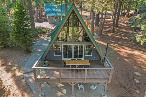 A-frame cabin surrounded by trees, featuring a deck with a table and chairs, and large windows reflecting the forest.