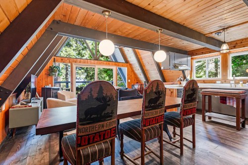 Cozy A-frame interior with wooden beams, a dining table, and chairs featuring moose designs, overlooking a scenic view.