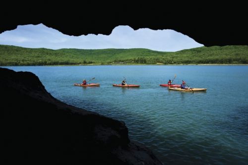 Four kayakers paddle on a calm lake, framed by a cave opening and surrounded by lush green hills.