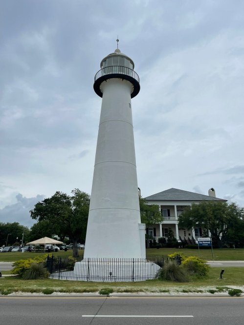 A tall white lighthouse stands beside a road, with a historic building and trees in the background under a cloudy sky.