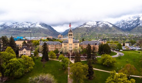Aerial view of a historic building surrounded by green trees and mountains, with snow-capped peaks in the background.