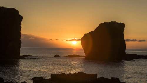 Sunset over the ocean, framed by two rocky formations silhouetted against a colorful sky.