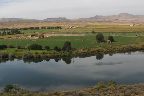 A serene landscape featuring a river reflecting green fields and distant mountains under a cloudy sky.