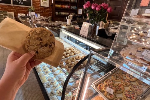 A hand holds a large cookie in front of a display case filled with various baked goods and a vase of roses.
