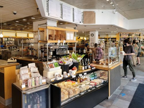 A cozy café interior with a display of baked goods, drinks, and flowers, featuring customers and staff in a welcoming atmosphere.