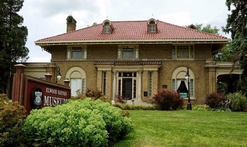 Historic brick mansion with a red tile roof, surrounded by greenery and a sign for the Elwood Haynes Museum.