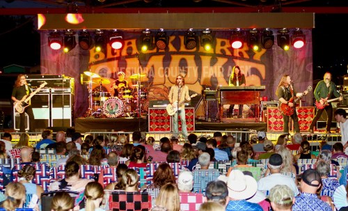 A band performs on stage at a county fair, with a large audience seated in colorful chairs. Bright lights illuminate the scene.
