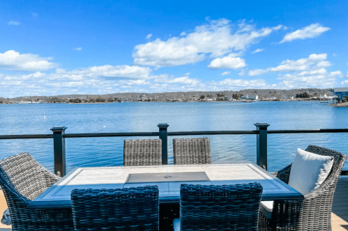 A scenic view of a lake from a deck with a table and chairs, under a bright blue sky with fluffy clouds.