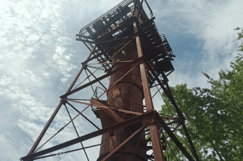 Rusty industrial tower against a cloudy sky, surrounded by greenery.