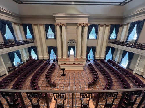 A spacious, elegant legislative chamber with rows of maroon chairs and large windows, featuring tall columns and blue drapes.