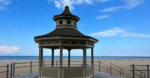 A gazebo on a sandy beach with a view of the ocean under a blue sky with scattered clouds.