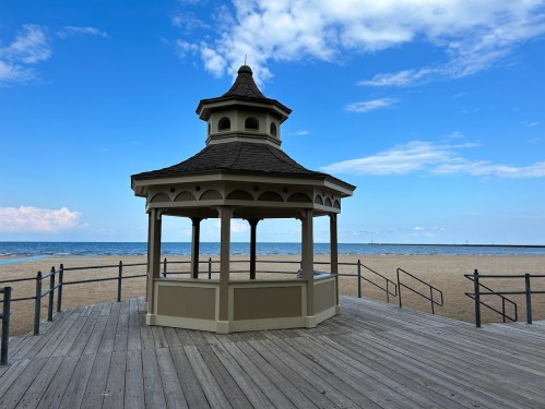 A gazebo on a wooden deck overlooking a sandy beach and calm ocean under a blue sky with scattered clouds.