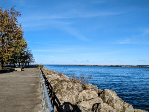 A serene lakeside view with a clear blue sky, rocky shoreline, and trees along a paved path.