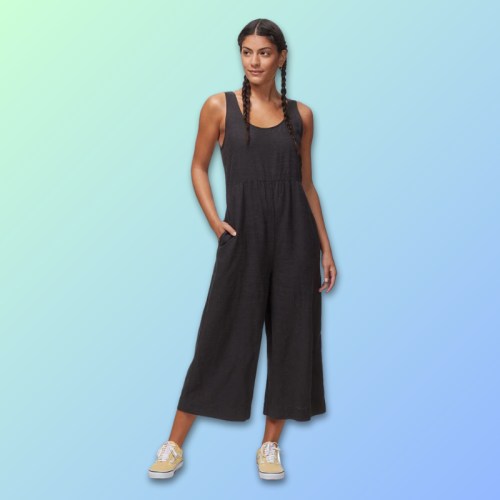 A woman in a black jumpsuit with pockets stands against a gradient background, wearing yellow sneakers and braided hair.