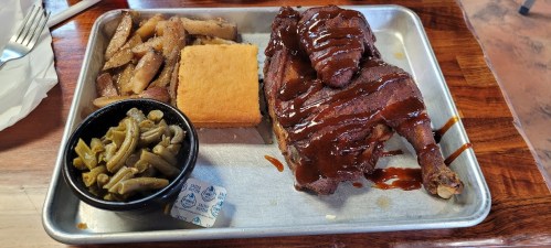 A metal tray with BBQ chicken, cornbread, green beans, and baked apples, drizzled with sauce.
