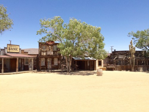 A deserted old western town with wooden buildings, a large tree, and a clear blue sky.