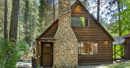 A cozy wooden cabin with a stone chimney, surrounded by tall trees and a serene creek.