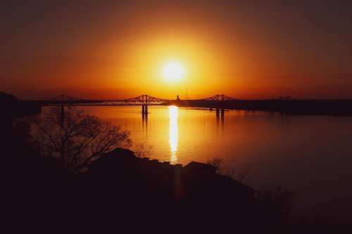 Sunset over a river, with a bridge silhouetted against the orange sky and reflections shimmering on the water.