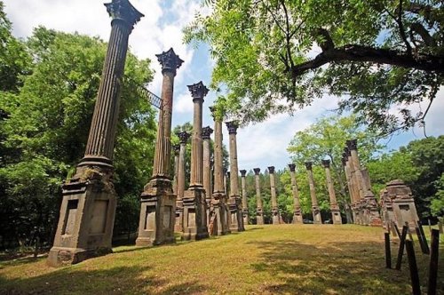 Ruins of tall columns surrounded by greenery under a blue sky, creating a serene and historical atmosphere.