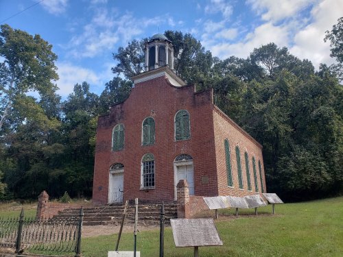Historic brick church with a bell tower, surrounded by trees and informational plaques in a grassy area.