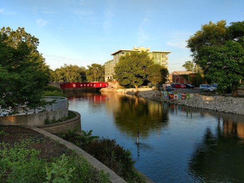 A serene river scene with trees, a red barge, and a modern building reflecting in the water under a clear blue sky.