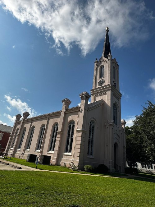 A pink church with a tall steeple and clock tower, set against a blue sky with scattered clouds. Green grass in the foreground.