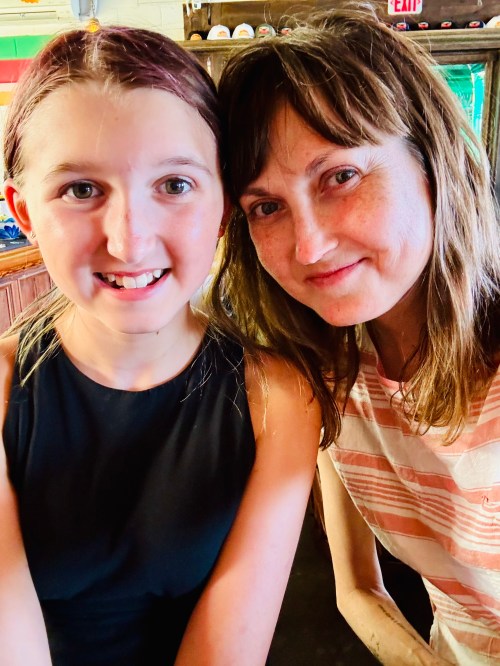 A smiling woman and a girl sit close together, both looking at the camera with warm expressions.
