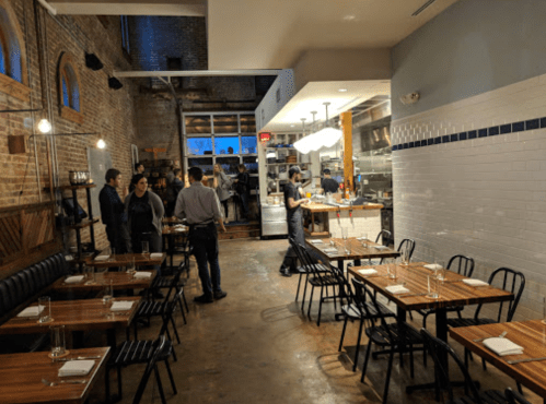 A cozy restaurant interior with wooden tables, black chairs, and a visible kitchen area, featuring diners and staff.