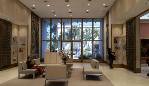 A spacious lobby with modern seating, large windows, and palm trees visible outside. People are engaged in various activities.