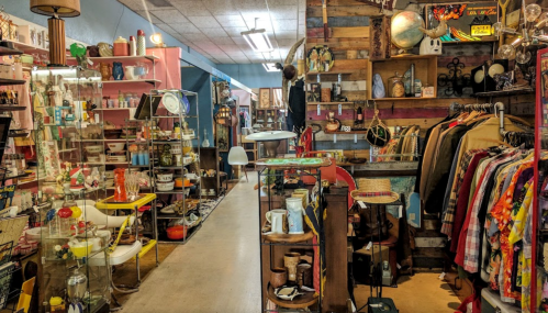 A vibrant vintage shop filled with eclectic items, clothing racks, and colorful displays along a narrow aisle.