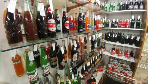 A display of various vintage soda bottles on glass shelves, showcasing colorful labels and different brands.