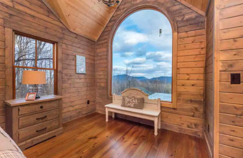 Cozy wooden room with large arched window showcasing mountain views and a bench by the window.