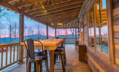 A rustic wooden porch with a large table and chairs, overlooking a scenic sunset and mountains in the background.