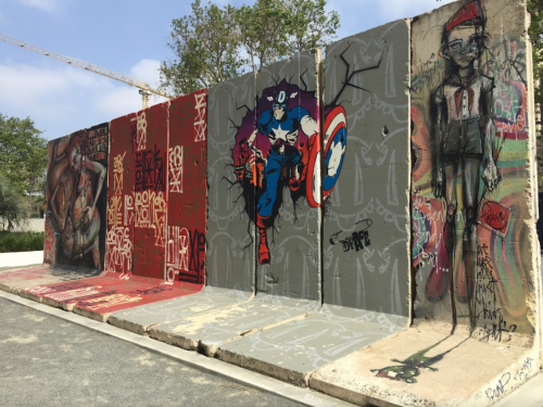 A colorful mural on concrete barriers featuring superhero and abstract art, surrounded by trees and a clear sky.