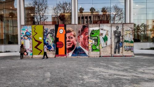 A mural featuring portraits of JFK and Ronald Reagan on a concrete wall, with people walking nearby.