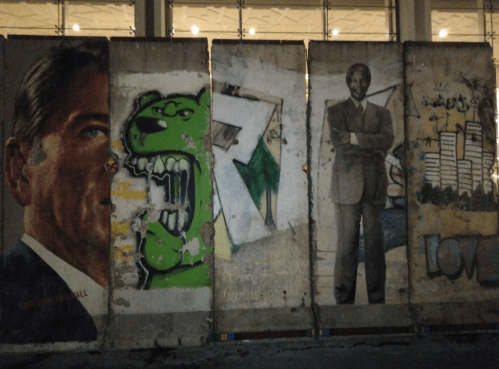 A mural on a concrete wall featuring a man's face, a green monster, and a suited figure, with urban art elements.