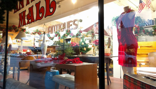 A festive store window display featuring holiday decorations, a red dress on a mannequin, and various home goods.