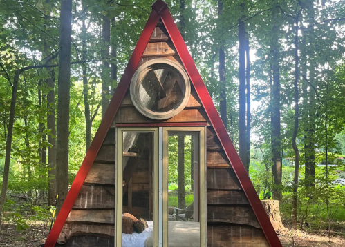 A small, triangular wooden cabin with a red roof, surrounded by lush green trees in a forest setting.