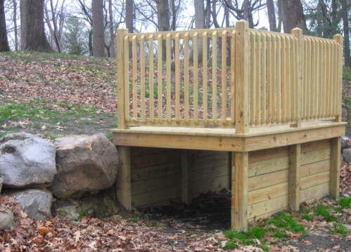 A wooden platform with a railing, elevated above ground, next to a rock formation and surrounded by fallen leaves.
