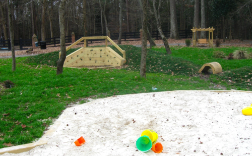 A playground scene featuring a sand area with colorful buckets, a climbing structure, and a small tunnel in a grassy setting.
