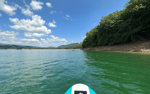 A paddleboarder glides on calm water, surrounded by green hills and a blue sky with fluffy clouds.
