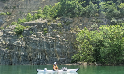 A person in a kayak paddles on calm water, surrounded by rocky cliffs and lush greenery.