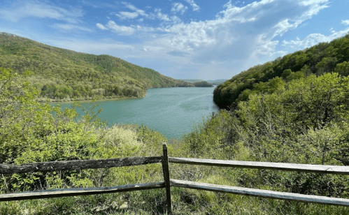 A serene lake surrounded by lush green hills under a blue sky with fluffy clouds, viewed from a wooden fence.