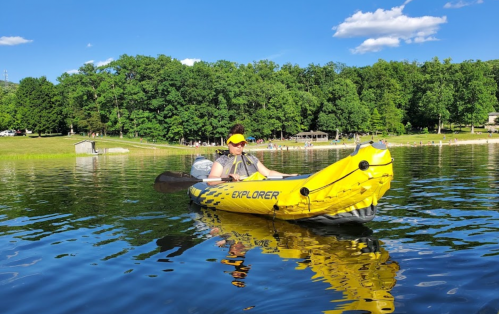 A person in a yellow kayak paddles on a calm lake surrounded by green trees and a sunny sky.