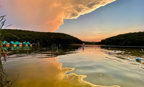 A serene lake at sunset, with colorful clouds reflecting on the water and lush green hills in the background.
